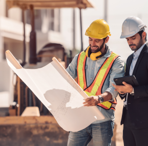 Two men in hard hats and safety vests examining a blueprint at a construction site.